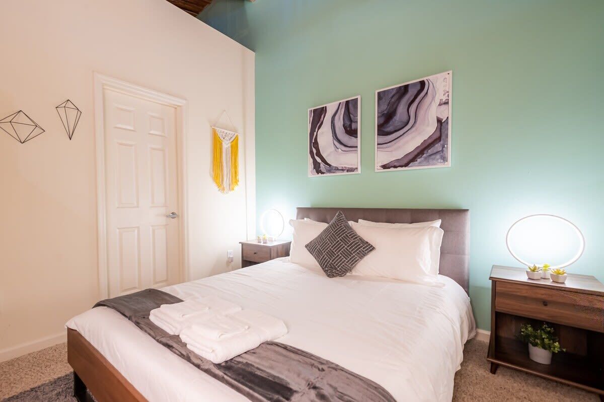A neatly made bed with a white comforter and a gray throw blanket, a wooden nightstand with a mirror and a small plant, and two abstract wall art pieces above the bed.