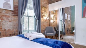Una camera d'albergo con una parete in mattoni, un letto grande con un tocco di blu, una poltrona grigia e uno specchio che riflette l'ambiente.