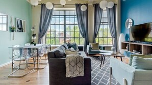 Un salon moderne avec un canapé, une table à manger et des chaises. De grandes fenêtres sont agrémentées de rideaux, et on y trouve également un téléviseur à écran plat et une bibliothèque.