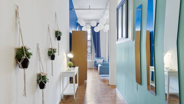 Un couloir doté d’un plancher en bois, de murs blancs et d’un mur d’accent bleu. On y trouve des suspensions en macramé avec des plantes vertes, une petite table d’appoint blanche avec une lampe, et un canapé bleu en arrière-plan.