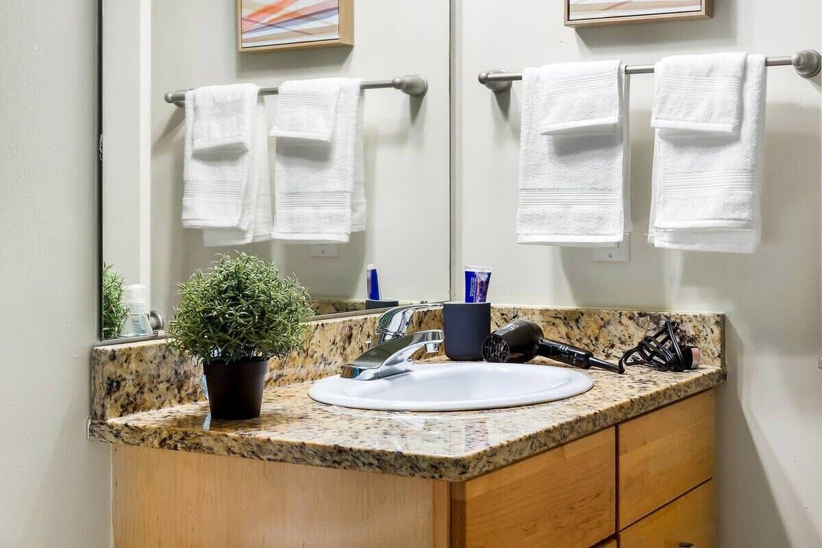 A bathroom with a granite countertop, a white sink, a potted plant, a mirror, and two towels hanging on a towel rack.