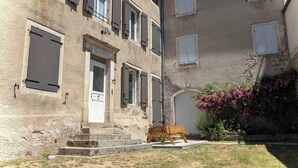 A stone building with a wooden bench, a small garden with pink flowers, and a white door with a glass panel. - L'oustal du Lac, à Proximité du lac de Naussac et des Sentiers de Randonnée (Auroux)