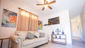 Una sala de estar con un sofá color beige, almohadones decorativos, una mesita de madera con jarrones y un ventilador de techo.