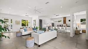 Amplia sala de estar con sofá blanco, una mesita de centro y ventilador de techo. Zona de cocina con electrodomésticos modernos y taburetes de bar.