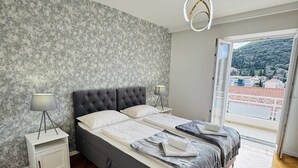 A modern bedroom with a grey tufted bed, a white bedside table, a fluffy grey rug, and a view of a hillside through a large window. - KABLAR'S SUNNY SUITES 2BR apartment with balcony (Dubrovnik)