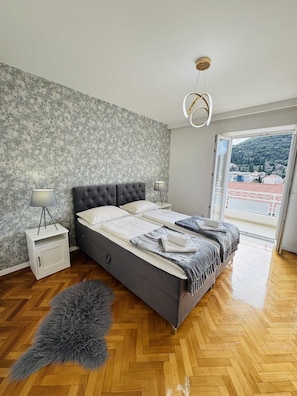 A modern bedroom with a grey tufted bed, a white bedside table, a fluffy grey rug, and a view of a hillside through a large window.