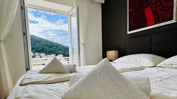 A hotel room with a bed, white linens, a view of the beach, and a red abstract painting on the wall. - KABLAR'S SUNNY SUITES 2BR apartment with balcony (Dubrovnik)