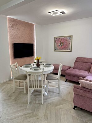 Un comedor con una mesa redonda preparada para cuatro personas, sillas blancas, un sofá rosa, un televisor de pantalla plana montado en la pared y un cuadro abstracto enmarcado.
