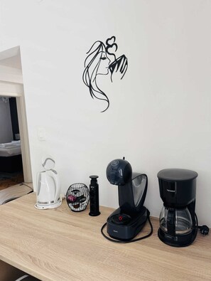 Una habitación con una pared blanca, un adhesivo mural negro de una mujer, un hervidor blanco, un hervidor eléctrico negro, una cafetera negra y otro hervidor eléctrico negro sobre una mesa de madera.