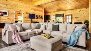 A cozy log cabin interior with a beige sectional sofa, a wooden coffee table with fruit, and a kitchen area visible in the background.