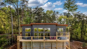 A modern two-story house with large windows and a balcony, surrounded by a forest.
