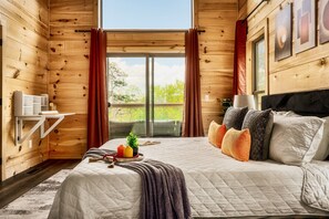 Une chambre dans une cabane en bois, avec un grand lit, une petite table ornée d’une plante et une vue dégagée sur l’extérieur grâce à une porte coulissante.