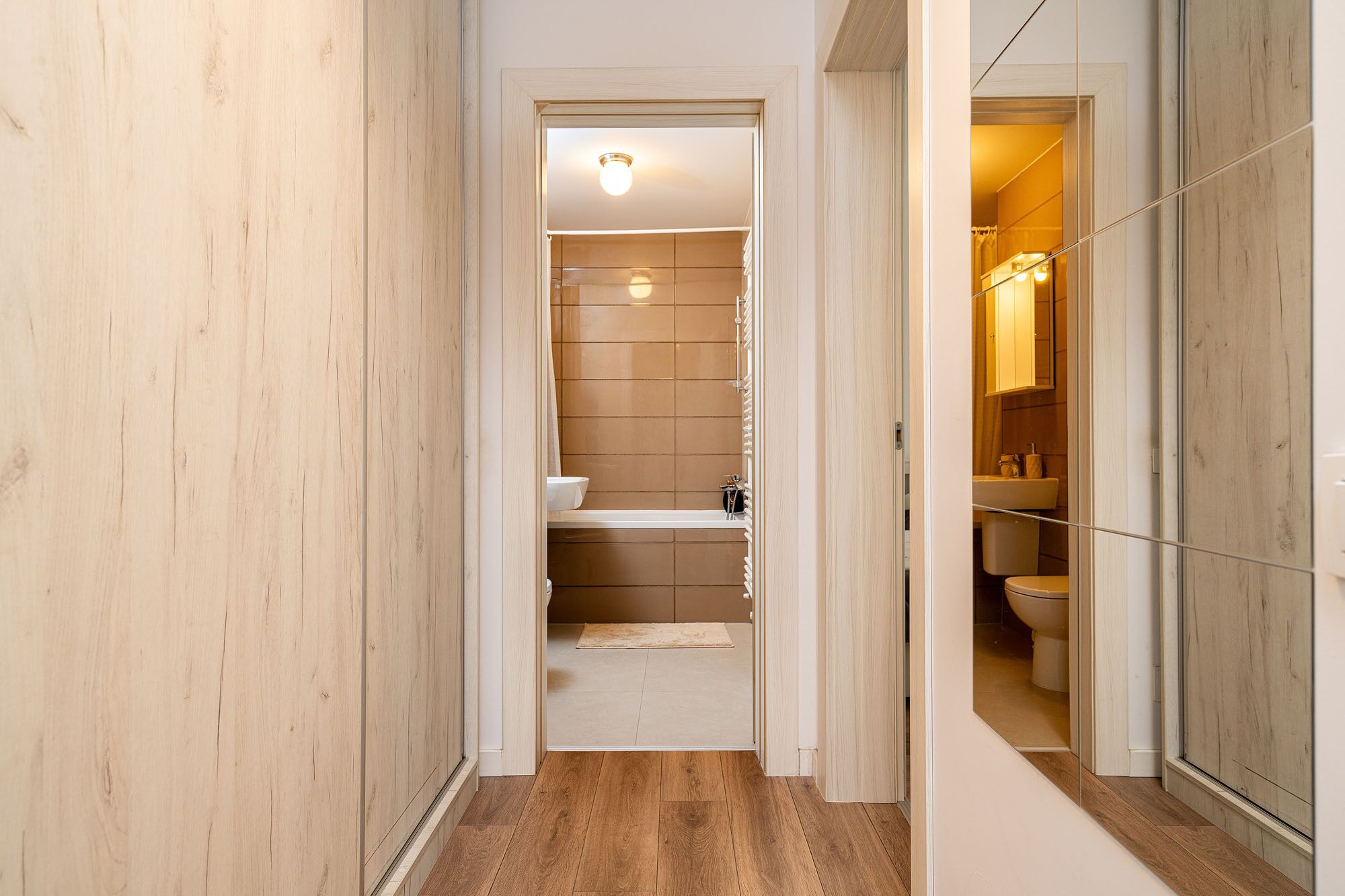 Ein schmaler Korridor mit Holzdielen und Wänden, der zu einem Badezimmer mit Toilette, Waschbecken und Duschbereich führt.