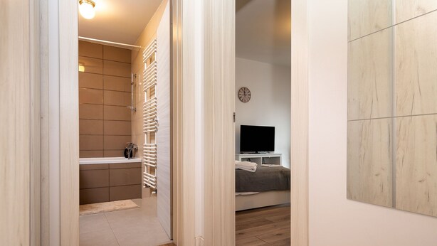 Un couloir mène à une salle de bain équipée d’une douche, de toilettes et d’un lavabo. La pièce adjacente contient un lit et un téléviseur.