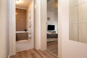 A hallway leading to a bathroom with a shower, a toilet, and a sink. There is a bed with a TV in the adjacent room.