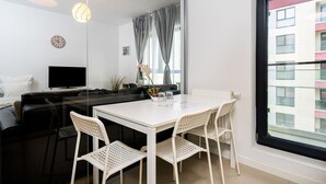 Un salon moderne avec un canapé blanc, un téléviseur, une table à manger avec des chaises et une grande fenêtre avec des rideaux.
