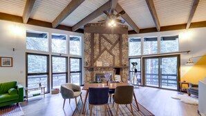 A spacious living room with a stone fireplace, large windows, and a high ceiling with exposed beams.
