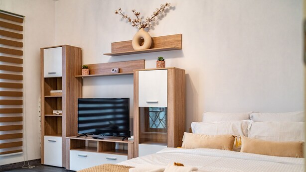 Un dormitorio moderno con un televisor sobre un mueble bajo de madera, una cama con ropa de cama blanca y un jarrón decorativo en una estantería.