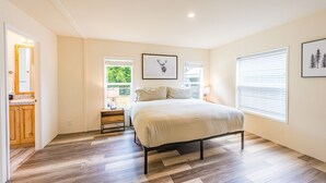 Une chambre à coucher comprenant un lit, une table de chevet, une fenêtre avec des stores et une porte donnant sur une salle de bain.