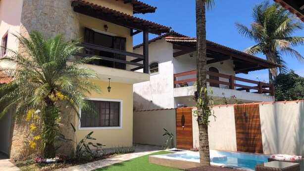 A two-story house with a stone pillar, a balcony, a swimming pool, and a well-maintained garden with palm trees. - Casa de Praia com Piscina Aquecida, ar Condicionado, Cervejeira e Bilhar!!! (Vista Linda Bertioga)
