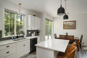 A modern kitchen with white cabinetry, a central island, and a dining area with a table and chairs. - Luxury Cabin with Hot Tub, Fire Pit, Fireplace (Rhododendron)