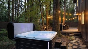 A hot tub in a forested backyard with string lights, a deck, and a wooden fence.