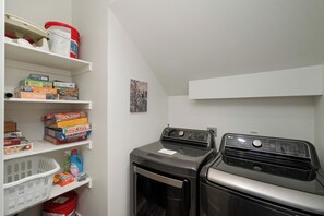 Un lavadero con lavadora, secadora, estantes con juegos de mesa y una pequeña foto en la pared.