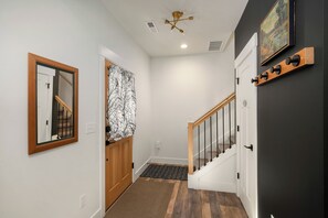 Un pasillo con puerta de madera, espejo, cortina estampada, escalera con barandilla de madera y una lámpara fijada a la pared.