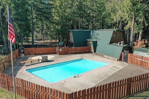 A rectangular swimming pool surrounded by a wooden fence, with a flagpole and a covered structure in the background. - Luxury Cabin with Hot Tub, Fire Pit, Fireplace (Rhododendron)