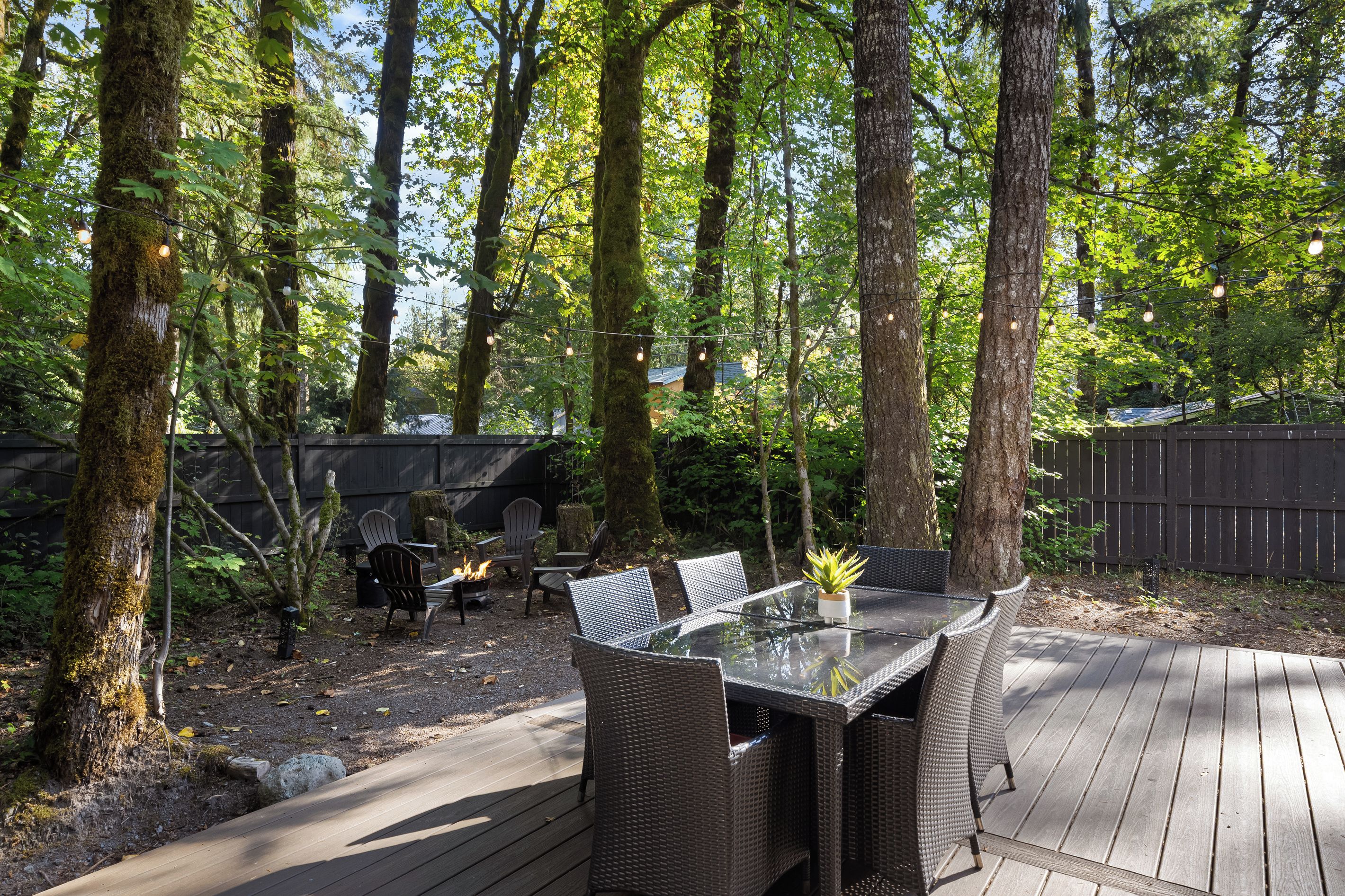 An outdoor dining area with a glass table and wicker chairs, surrounded by trees and a wooden deck.