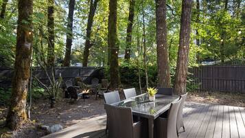 An outdoor dining area with a glass table and wicker chairs, surrounded by trees and a wooden deck.