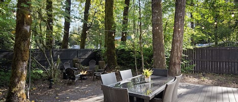 Un espace extérieur aménagé pour les repas, avec une table en verre et des chaises en osier, entouré d’arbres et doté d’une terrasse en bois.