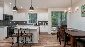 A modern kitchen with white cabinets, a central island, black pendant lighting, and a dining area with wooden furniture.