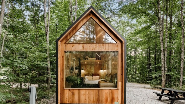 Una cabaña de madera con techo triangular, ventanas grandes y una cama visible en su interior.