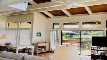 A spacious living room with a high ceiling, large windows, a ceiling fan, and a television.