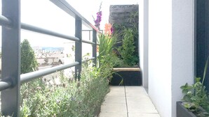 Un balcón estrecho con barandilla de metal, piso de concreto y vista a un paisaje urbano al fondo.
