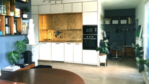 Una cocina moderna con armarios blancos, un horno negro y una encimera de madera. Hay plantas en los estantes y una silla en la zona de comedor.