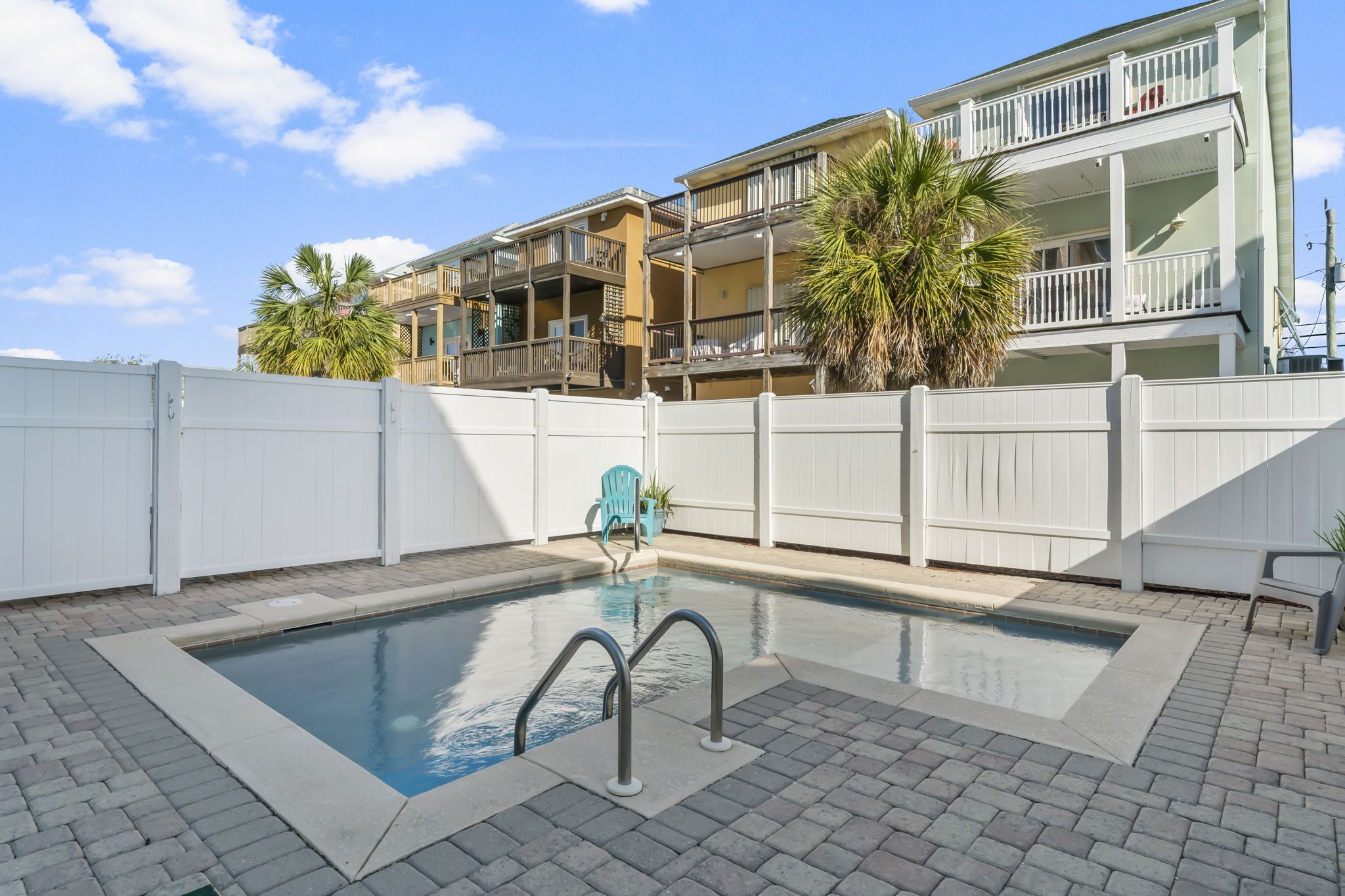 A swimming pool with a white fence, a blue chair, and a multi-story building in the background.