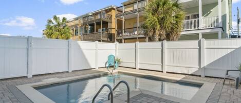 A swimming pool with a white fence, a blue chair, and a multi-story building in the background.