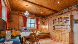 A cozy cabin interior with a wooden dining table, chairs, a bench with cushions, a kitchen area with cabinets, and a hanging light fixture. - Ferienhaus Taubenstein by Interhome (Neukirchen am Großvenediger)