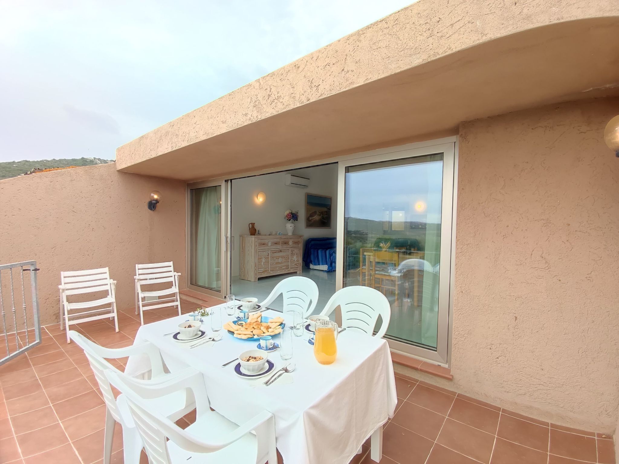 A terrace with a table set for dining, chairs, and a sliding glass door leading to an indoor area with a sofa and a table.
