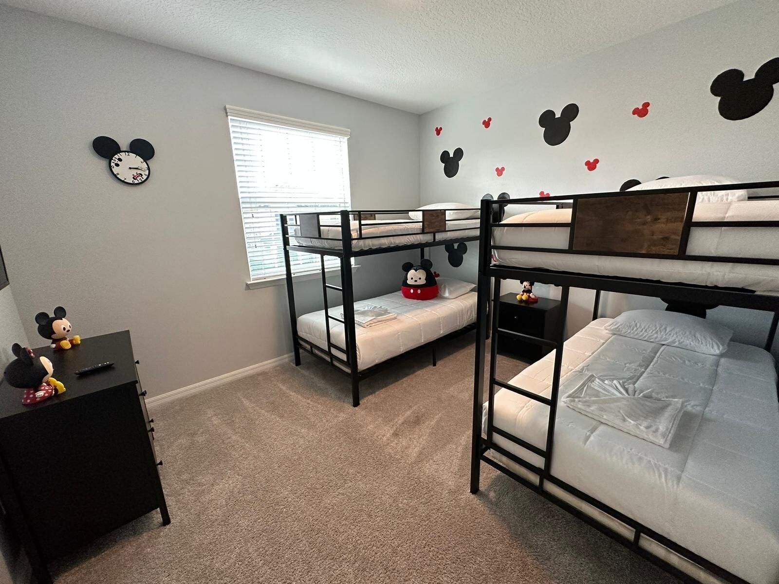Une chambre avec des lits superposés, un réveil à l’effigie de Mickey Mouse accroché au mur, un petit bureau orné d’une figurine de Mickey Mouse et une fenêtre avec des stores.