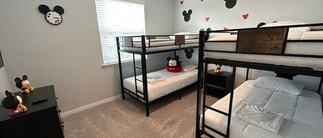 Une chambre avec des lits superposés, un réveil à l’effigie de Mickey Mouse accroché au mur, un petit bureau orné d’une figurine de Mickey Mouse et une fenêtre avec des stores.