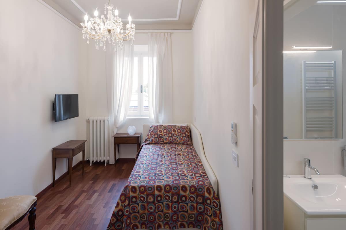 Une chambre à coucher comprenant un lit, une table de chevet, une chaise, un téléviseur, un lustre, une fenêtre avec des rideaux, un radiateur, et une salle de bain avec un lavabo et un miroir.