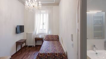 Ein Schlafzimmer mit Bett, Nachttisch, Sessel, Fernseher, einer Kristallleuchtre, einem Fenster mit Vorhängen, einer Heizung sowie einem Badezimmer mit Waschbecken und Spiegel.