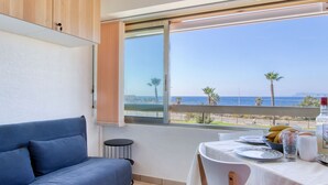 Ein modernes Zimmer mit einem blauen Sofa, einem Esstisch mit Tellern und einer Flasche, und einem großen Fenster mit Blick auf das Meer und Palmen.