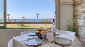 Ein Esstisch mit Tellern, einem Glas, einer Flasche und einer Schale mit Obst an einem Fenster mit Blick auf das Meer und Palmen.