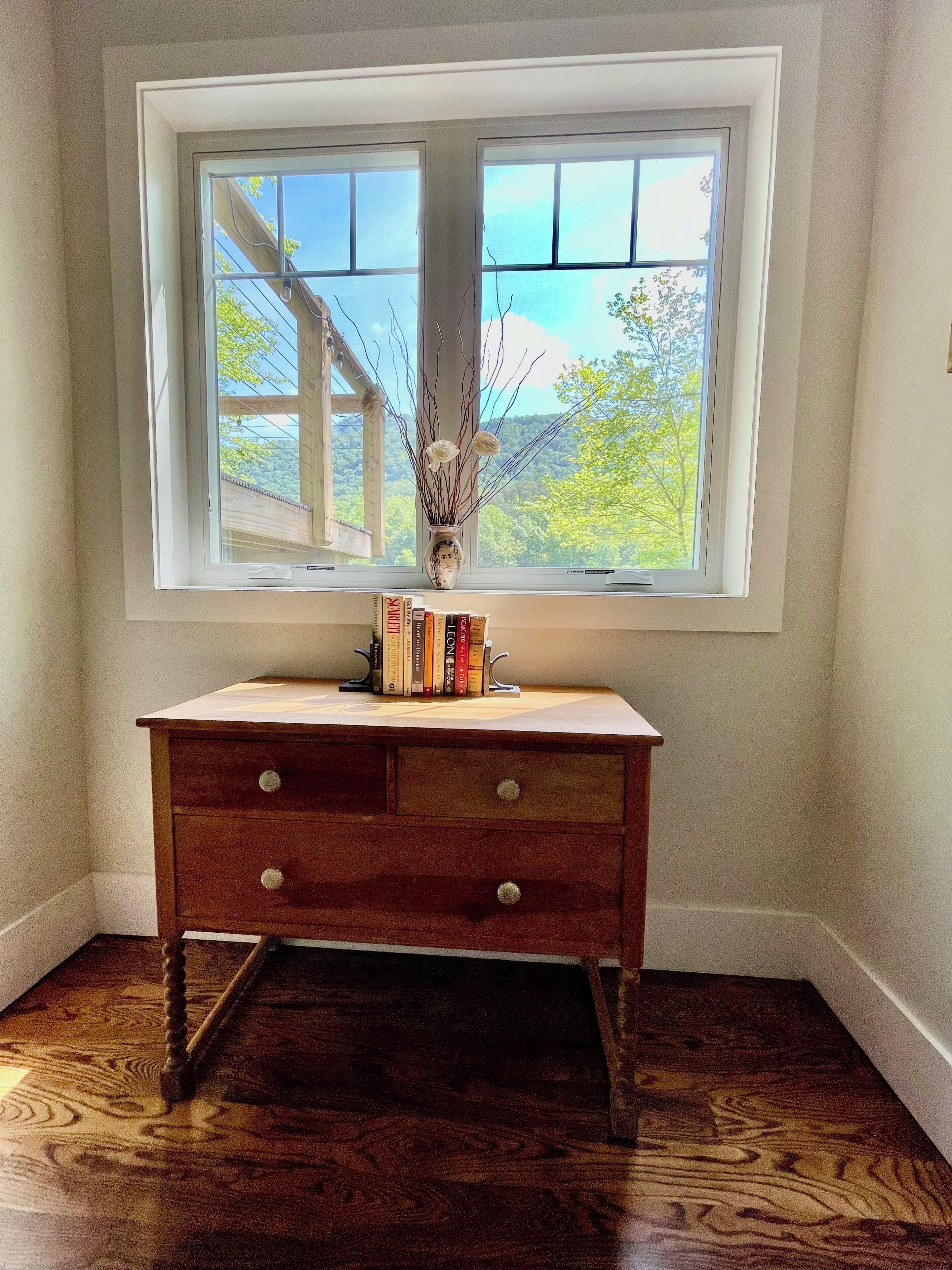 A corner room with a wooden desk, a window with a view of trees, and a vase with branches.