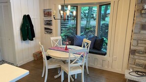 En matplats med ett runt bord, vita stolar och en bänk med kuddar. Det finns ett fönster med utsikt över träd och en stenmur.
