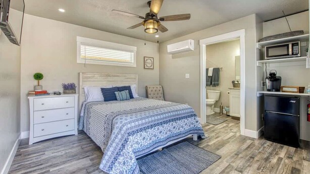 A bedroom with a bed, a nightstand, a dresser, a ceiling fan, and a kitchenette area.